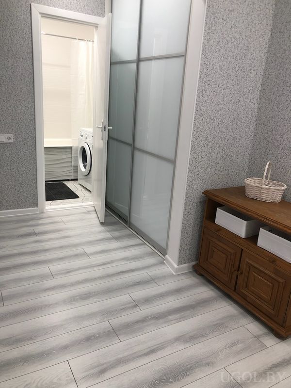 Проход с плиткой, белой дверью, шкафом с матовыми панелями и видом на ванную с чёрным ковриком.