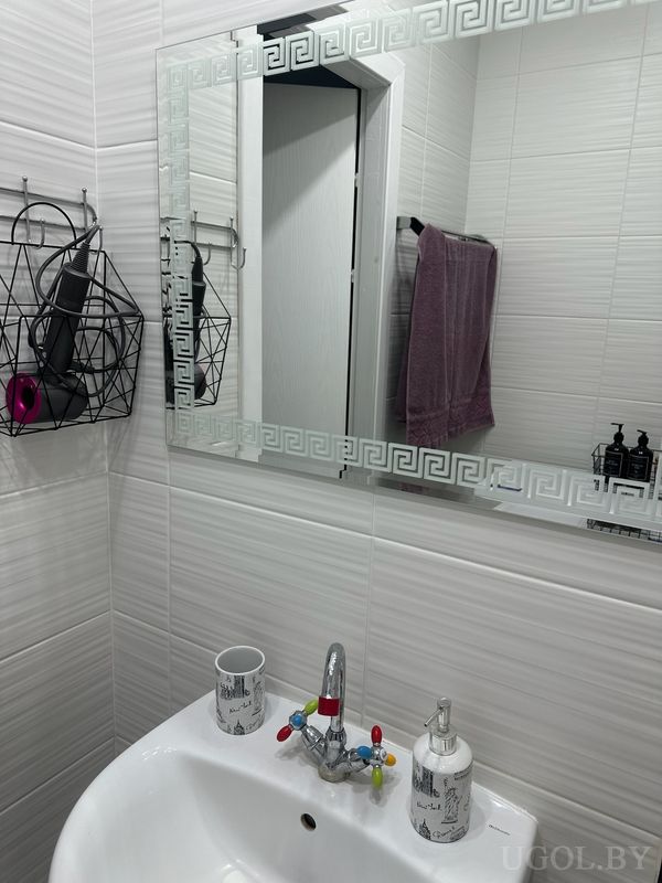 Комната с умывальником, зеркалом с греческим орнаментом, дозатором, полотенцем и корзиной с феном.