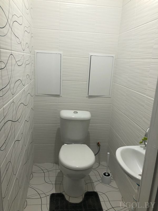 Узкое помещение с унитазом, умывальником, ёршиком, декоративной плиткой и ковриком с серым узором.