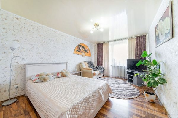 Комната с кроватью, бежевым покрывалом, игрушками, креслом, телевизором, растением и ковром у окна.