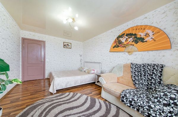 Интерьер с кроватью, диваном с пледом, декоративным веером, растением, ковром и светильником на потолке.