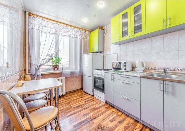 Интерьер с плитой, холодильником, микроволновкой, столом у окна и шторами с растением на подоконнике.