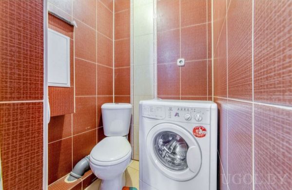 Компактное помещение с унитазом, машиной, шкафчиком, трубами и плиткой в красно-белой гамме.