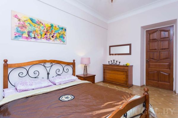 Уютная спальня с деревянной кроватью и металлическим изголовьем, коричневым покрывалом, сиреневыми подушками и яркой абстрактной картиной над кроватью.