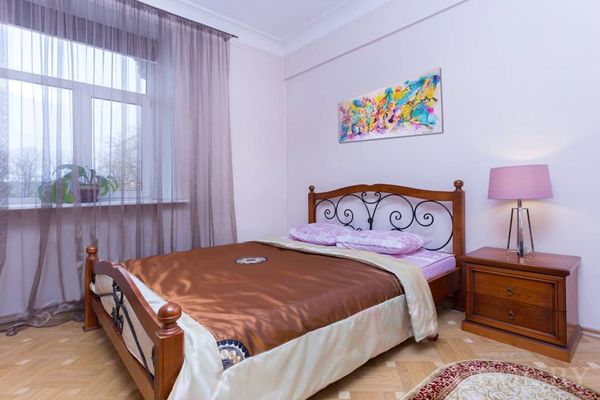 Светлая спальня с деревянной кроватью и металлическим изголовьем, коричневым покрывалом, сиреневыми подушками и большим окном с коричневыми шторами.