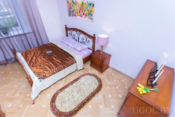 Спальня с кроватью и металлическим изголовьем, сиреневыми подушками, коричневым покрывалом, ковром с узором и тюльпанами на комоде.