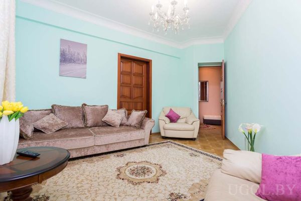 Светлая гостиная с мятными стенами, бежевым диваном, креслом, круглым столиком с тюльпанами и большим ковром с узором.