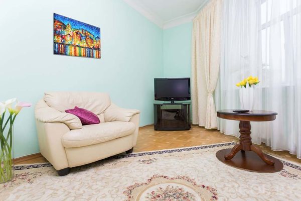 Уютная гостиная с кремовым креслом, круглым столиком с жёлтыми цветами, телевизором и картиной на стене.