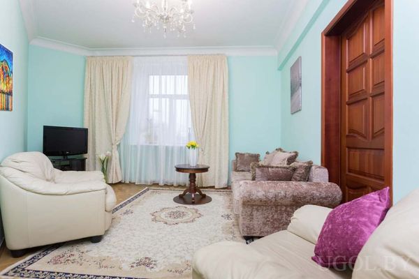 Светлая гостиная с сиреневым диваном, двумя креслами, круглым столиком с цветами и телевизором у окна.