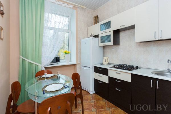 Компактная кухня с белыми и коричневыми шкафами, плитой, холодильником и стеклянным столом с сервировкой.