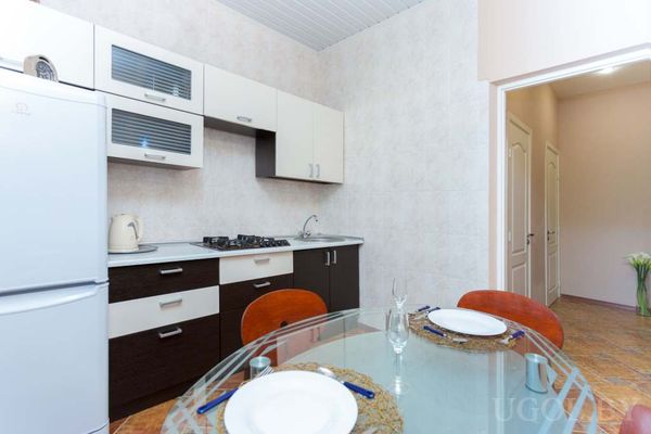 Современная кухня с белыми и коричневыми шкафами, плитой, холодильником и стеклянным столом с оранжевыми стульями.