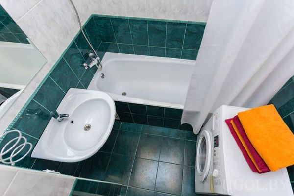 Компактная ванная с зелёной плиткой, ванной, раковиной, стиральной машиной и стопкой ярких полотенец.