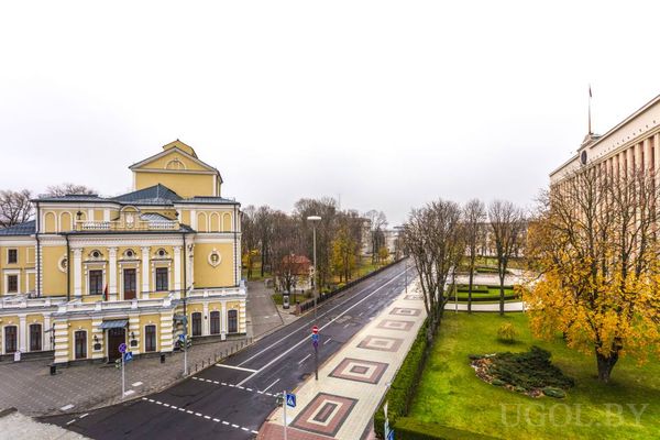 Городская улица с жёлтым зданием театра, административным зданием с флагом и осенними деревьями.