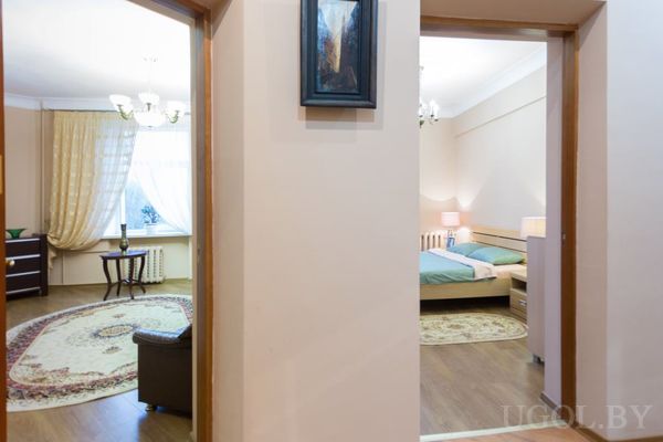 Интерьер с видом из коридора: слева гостиная с круглым ковром и столиком у окна, справа спальня с зелёным покрывалом.