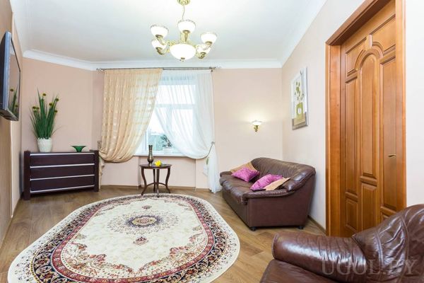 Комната с круглым ковром, коричневым диваном с подушками, круглым столиком у окна и телевизором на стене.