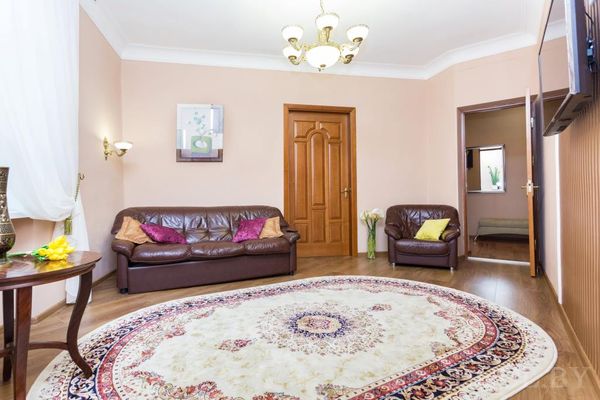 Уютная гостиная с круглым ковром, диваном и креслом с подушками, дверью в спальню и телевизором на стене.