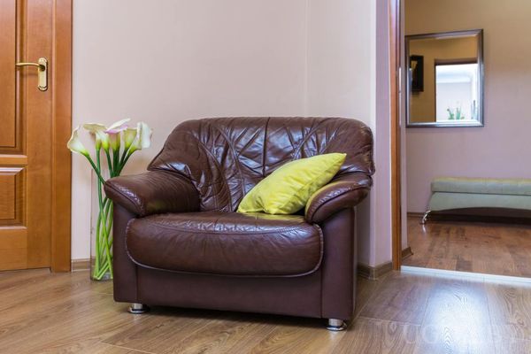 Интерьерный уголок с коричневым креслом, зелёной подушкой, вазой с белыми каллами и дверью в соседнюю комнату.