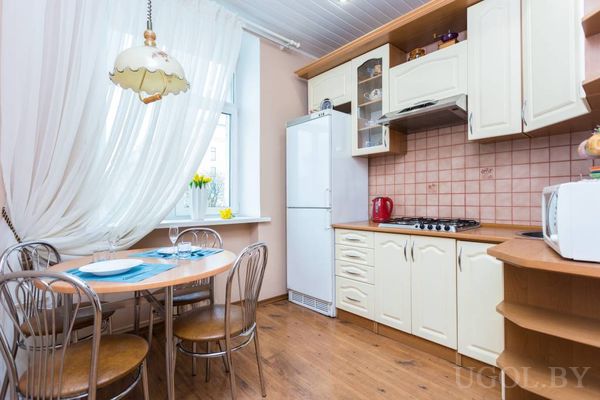 Светлая кухня с деревянным столом, сервированным посудой, белым холодильником, плитой и вазой с жёлтыми цветами.