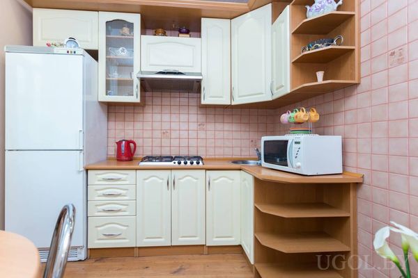 Небольшая кухня с белыми шкафами, плитой, мойкой, микроволновкой и розовой плиткой на фартуке.