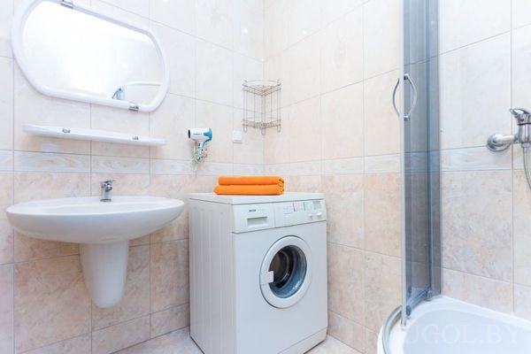 Небольшая ванная с душевой кабиной, стиральной машиной с оранжевым полотенцем и настенным феном.
