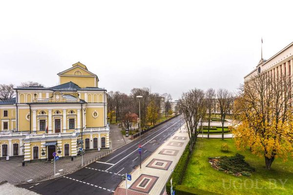 Широкая улица с жёлтым театром слева и административным зданием с флагом справа.