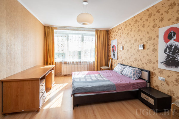 Кровать с узорным покрывалом, настенные светильники, картины, стол с полками и окно с белыми тюлевыми и жёлтыми шторами.