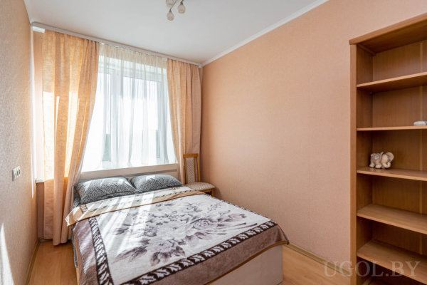 Кровать у окна с цветочным покрывалом, деревянный стул, высокий шкаф с игрушкой и мягкий свет.