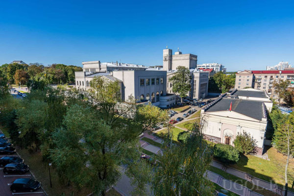 Классическое здание с башней, деревья, припаркованные машины и яркое дневное освещение на фоне голубого неба.