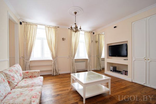 Элегантная комната с цветочным диваном, белым журнальным столиком и встроенной нишей с телевизором. Три высоких окна с бежевыми шторами пропускают мягкий дневной свет.