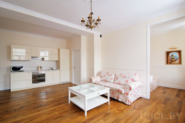 Открытое пространство с белой кухней, встроенной техникой и диваном напротив журнального столика. В нише — картина с кораблём и декоративная полка.