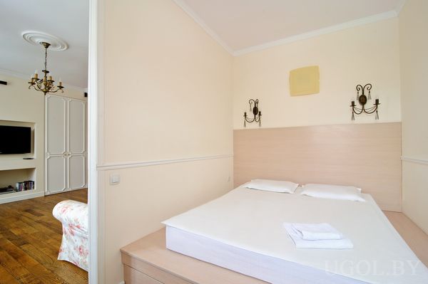 Кровать с белым бельём и светильниками на стене. Через дверной проём видна гостиная с телевизором, креслом и люстрой, создающая ощущение единого пространства.