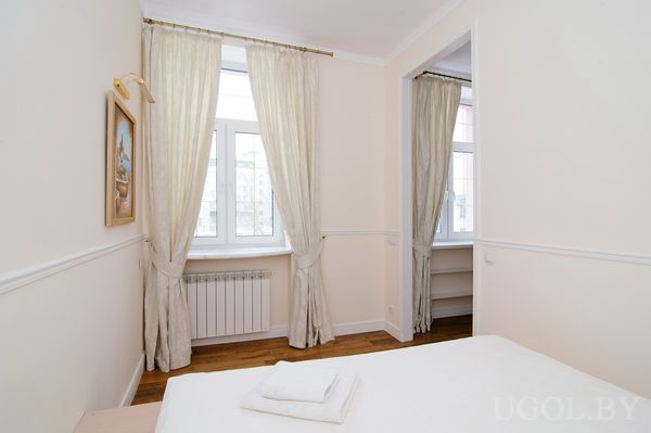 Небольшая комната с двумя окнами, белым бельём и встроенными полками. Между окнами — картина, подчеркивающая уют и чистоту интерьера.