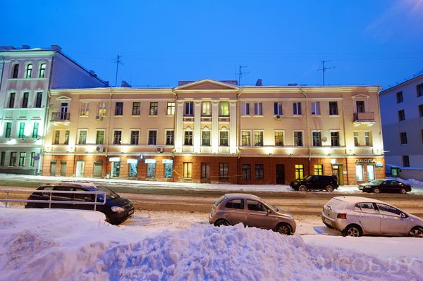 Трёхэтажное здание с подсвеченными витринами и снежной улицей. Тёплый свет окон контрастирует с холодным небом, создавая атмосферу зимнего города.