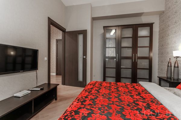 Интерьер спальни с кроватью в красно‑чёрном покрывале, тёмным шкафом с матовыми вставками, телевизором и прикроватной тумбой с вазами.