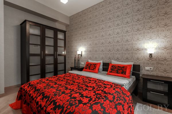 Элегантная спальня с серым изголовьем, красно‑чёрным покрывалом, настенными бра и большим шкафом с матовыми дверцами.
