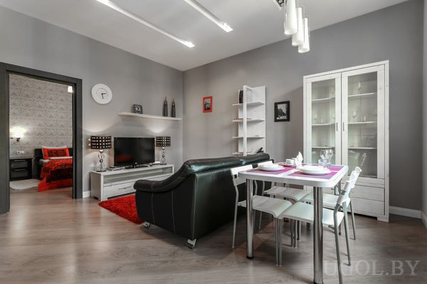 Современная гостиная с чёрным диваном, телевизором, красным ковром и обеденным столом на четыре персоны.