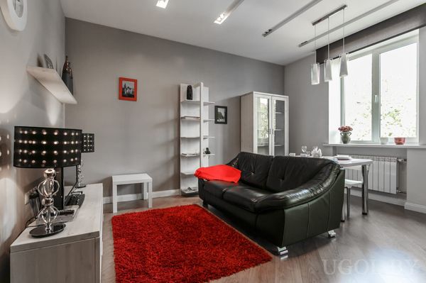Светлая гостиная с чёрным диваном, красным ковром, телевизором на тумбе и обеденной зоной у окна.