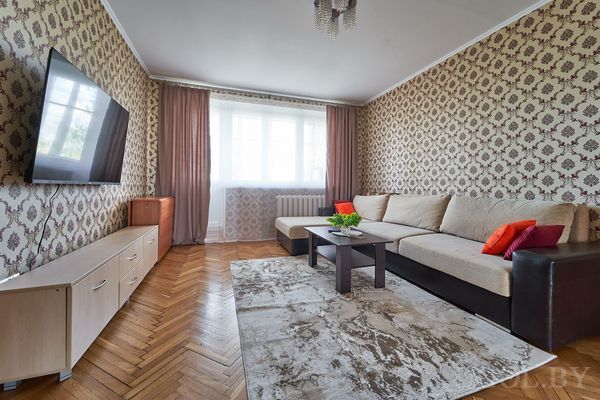 Современная гостиная с большим диваном, журнальным столиком, телевизором и окнами с тюлем и портьерами.