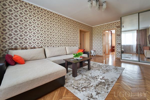 Комната с бежевым диваном, журнальным столиком, ковром и большим зеркальным шкафом на фоне обоев.