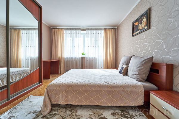 Уютная спальня с кроватью в бежевом текстиле, зеркальным шкафом и рабочим столом у окна.