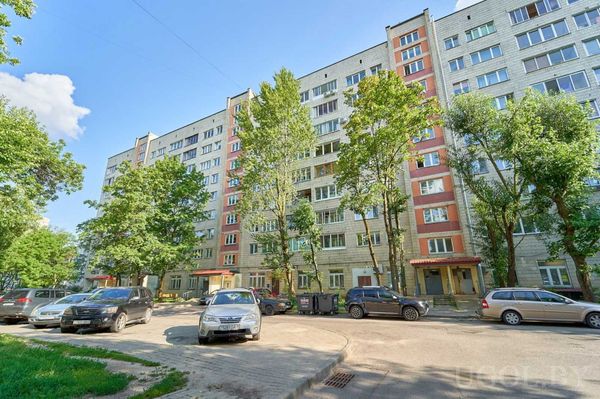 Многоэтажный дом с фасадом из светлого кирпича, красными акцентами и машинами у подъезда.