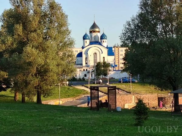 Вид на зелёную зону с деревьями, беседкой и православной церковью с куполами на фоне здания. На фото — архитектура, природа и люди в спокойной городской обстановке.