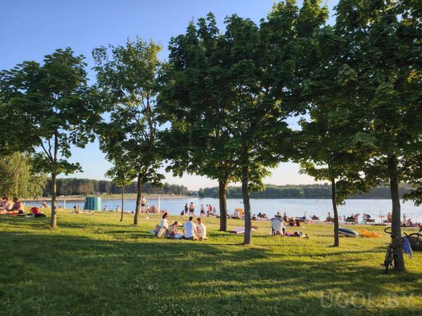 Летний парк с озером, деревьями и людьми на траве и песке. На фоне — лес и спокойная вода, в кадре — вечерний свет и тени.