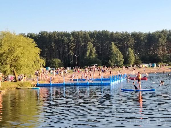 Оживлённый пляж с соснами, людьми в воде и на берегу, плавучим ограждением и активной атмосферой. Сцена — типичный летний день у озера.