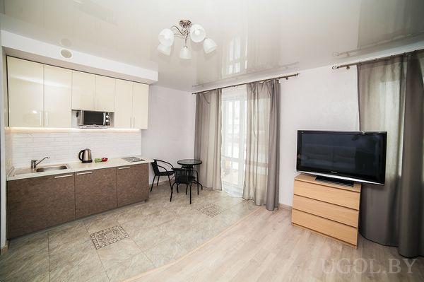 Современная студия с небольшой кухней, круглым столом на двоих и телевизором у окна с занавесками.