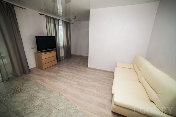 Светлая гостиная с кремовым кожаным диваном, телевизором на комоде и большими окнами с серыми шторами.