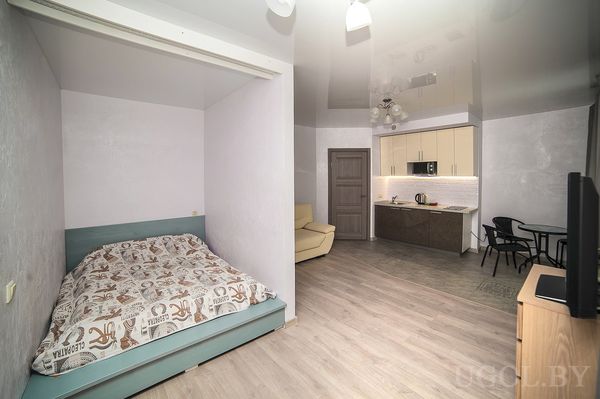 Студия с кроватью в нише, бежевым креслом, кухней с подсветкой и небольшим обеденным столом.