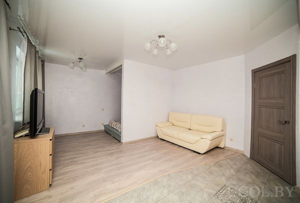 Комната с бежевым диваном, телевизором на тумбе и частично видимой кроватью в нише.