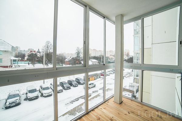 Застеклённый балкон с деревянным полом и видом на зимний городской двор с машинами и домами.