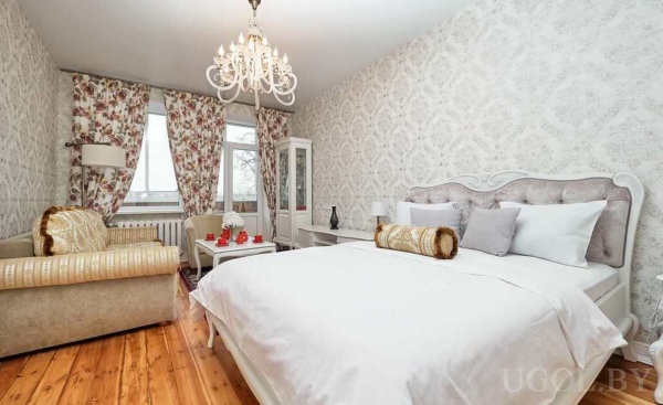 Элегантная спальня с большим белым ложем, бежевым диваном, люстрой и цветочными шторами.